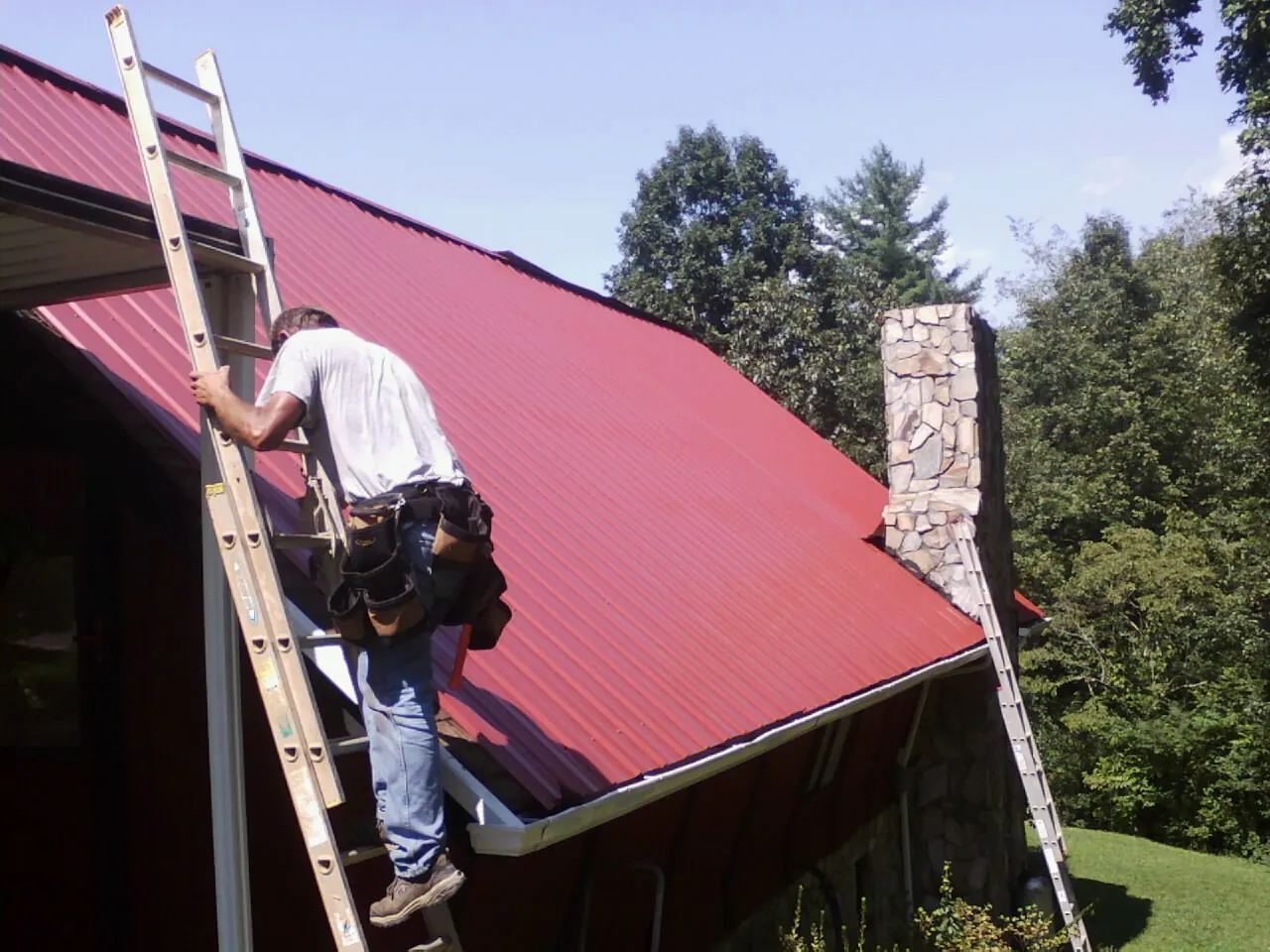 Roofer on a ladder beside a red metal roof and stone chimney — Soffit & Fascia Repair by Roofing Brothers in Welch